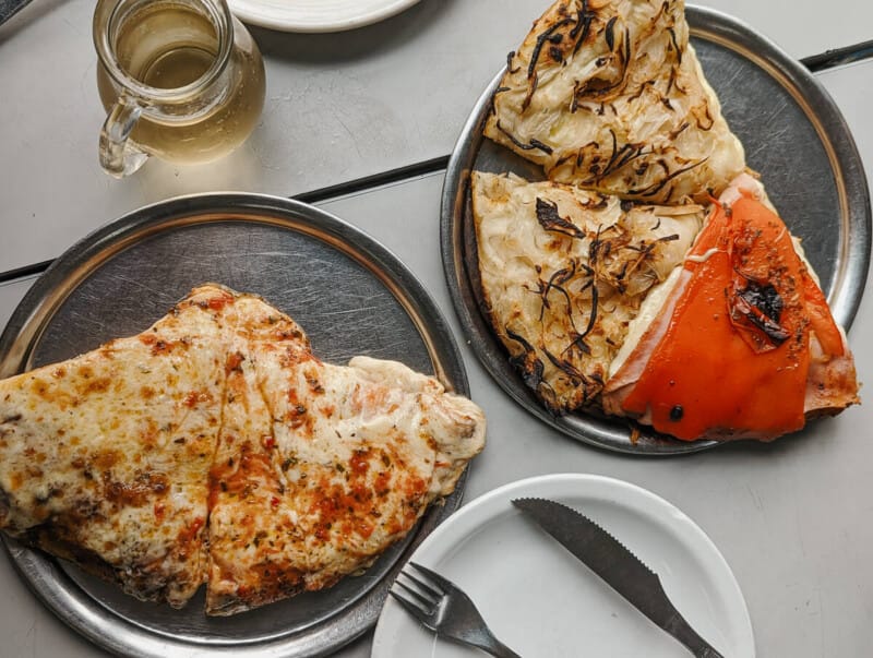 5 slices of pizza on two round metal dishes site on a white table next to a small jar of white wine