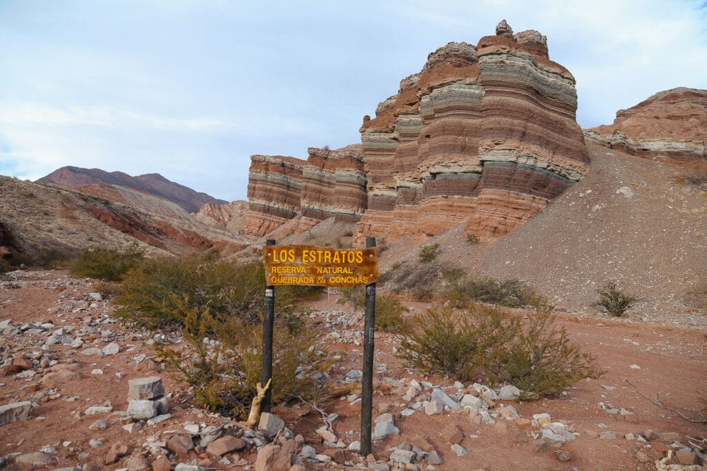 A sign marks the Los Estratos hike in the Quebrada de las Conchas in front of a rainbow colored mountain formation