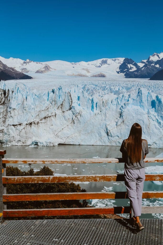 A woman leans against a handrail looking at a massive bright blue glacier