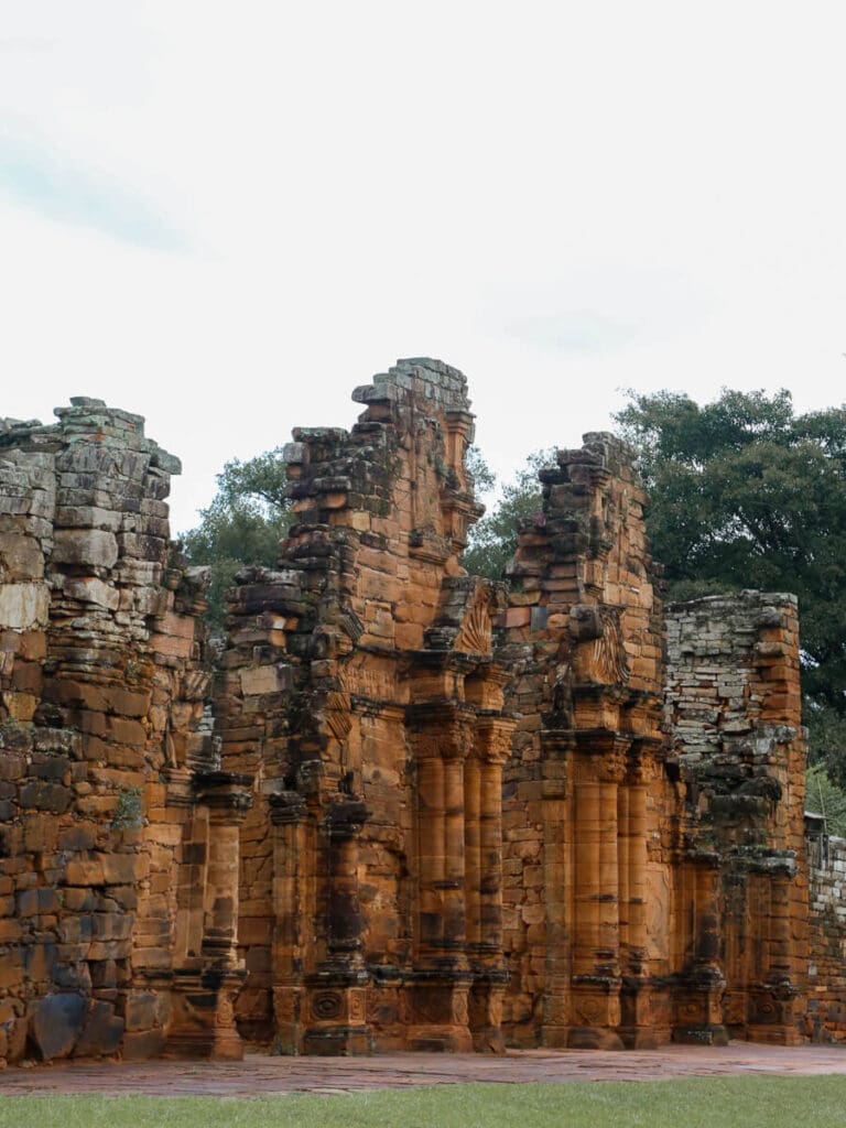 Red stone ruins of a Jesuit Mission stand on a green lawn
