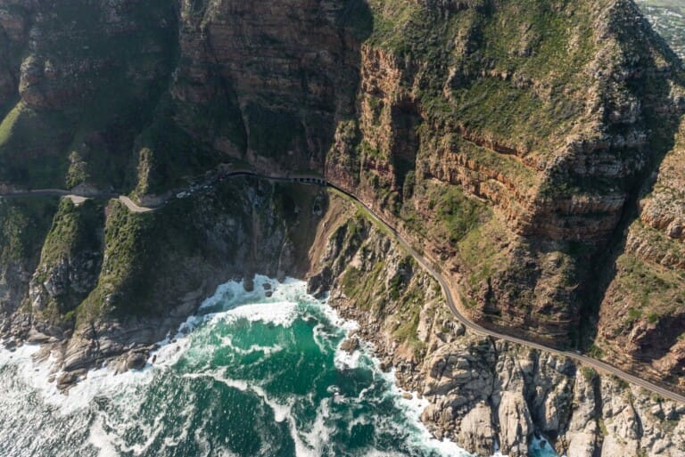 A road snakes along the rocky coast of a green mountain with the ocean below