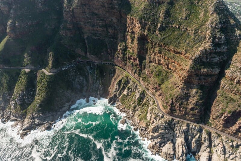 A road snakes along the rocky coast of a green mountain with the ocean below