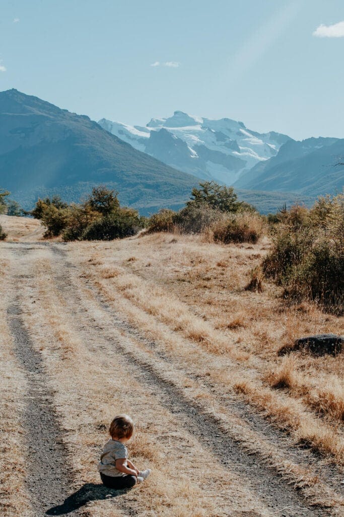 A baby is sitting with his back to the camera on a dirt road with mountains in the background