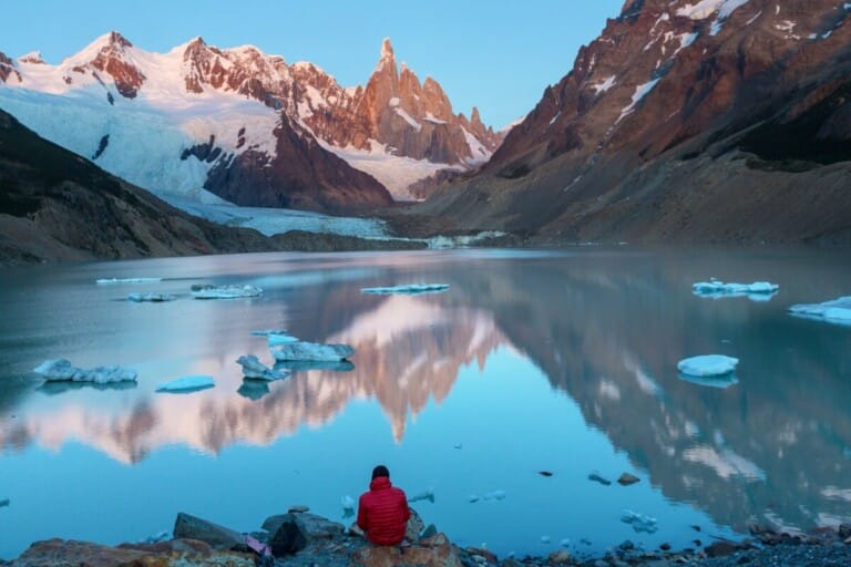 A man in a red coat sits by a lake at the mountains