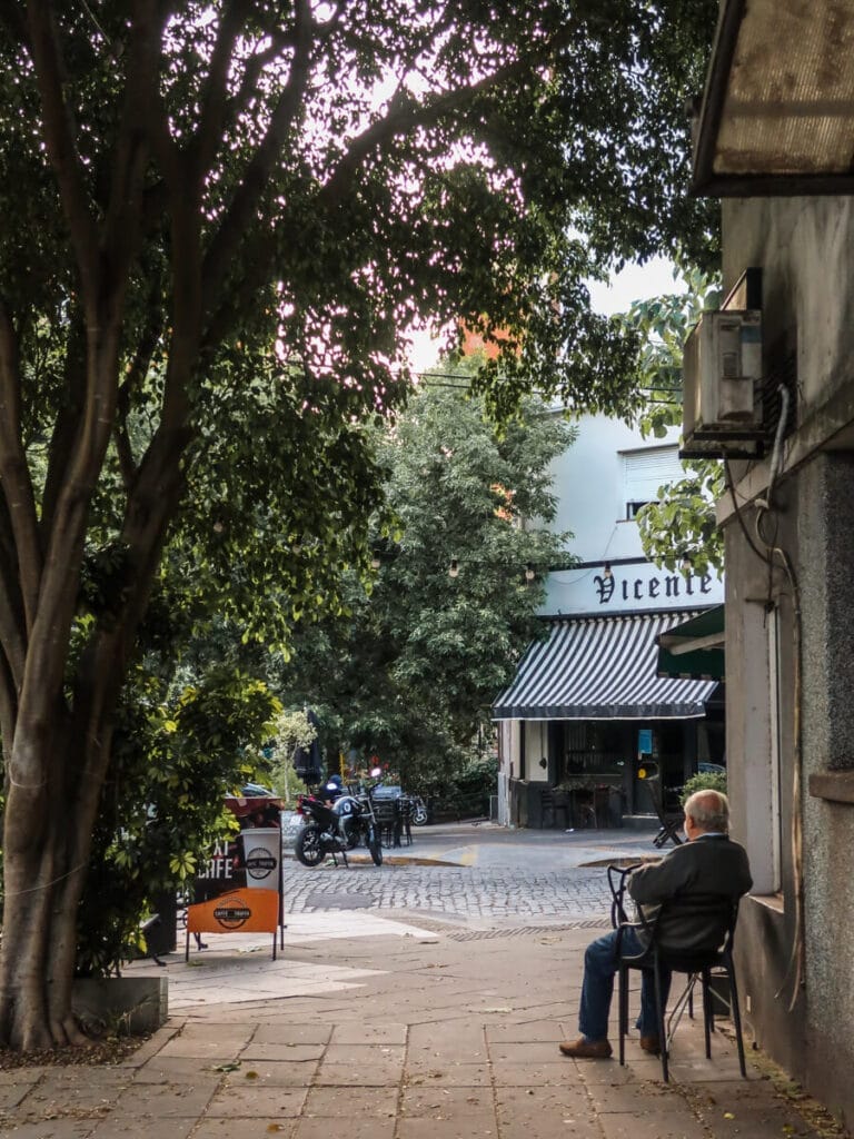 A man sitting in a chair on the sidewalk at a cafe under a canopy of trees.