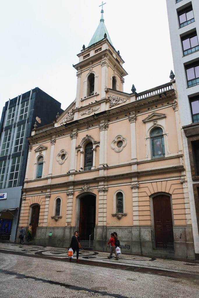 3 people walk down a cobblestone street in front of a pink two story church