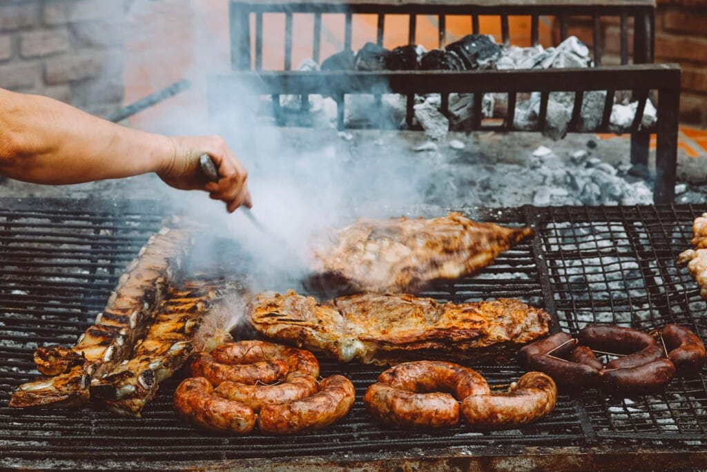 A hand holding a prong pokes at a steak on a grill filled with meat and sausage
