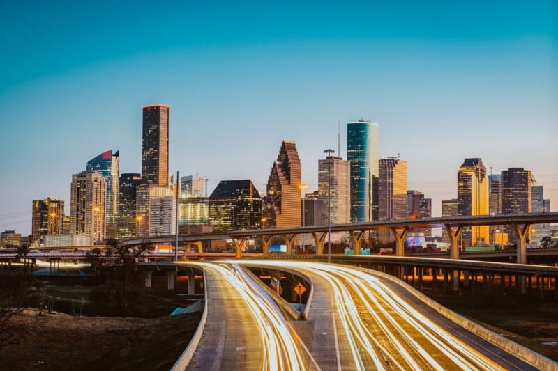 A highway with blurred tail lights in front of the skyline of Houston in the distance as the sunset