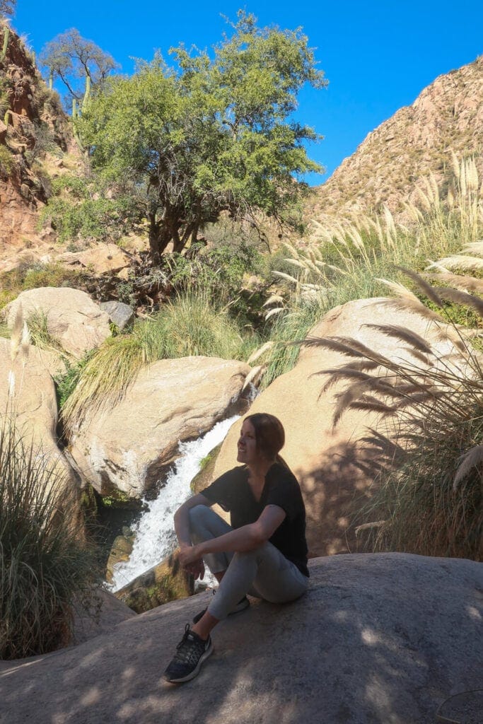 A woman sits on a boulder in front of a waterfall