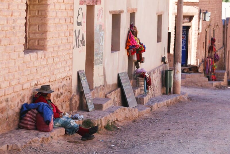 A woman sits on the side of a dirt road next to stucco buildings