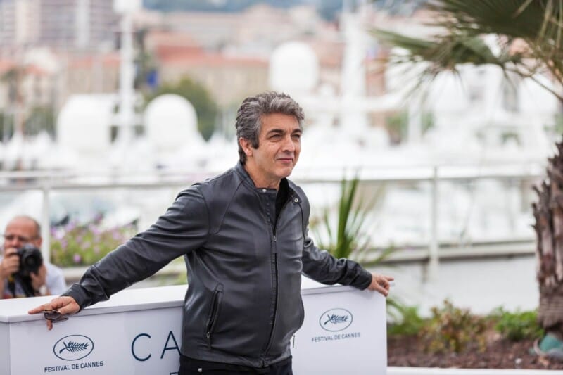 A man with grey hair and a leather jacket leans against a white podium