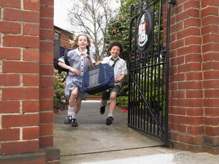 Two school children in private school uniforms run out of a gate to leave school