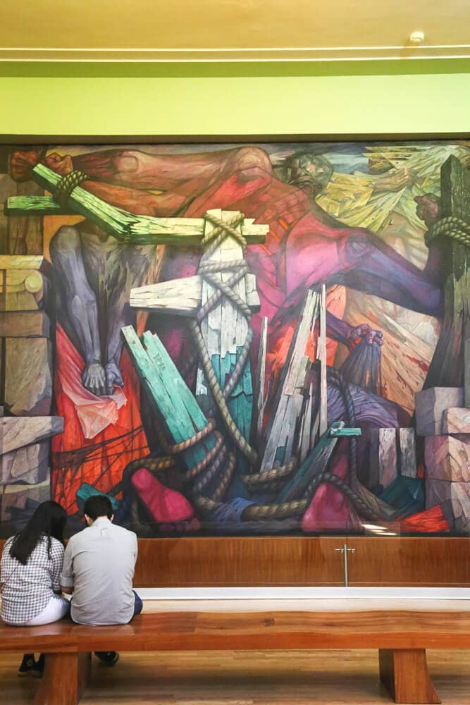 Two people sit on a bench in front of a Mural