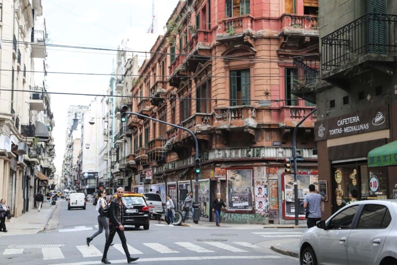 A man in a leather coat crosses the street in front of a run down red building
