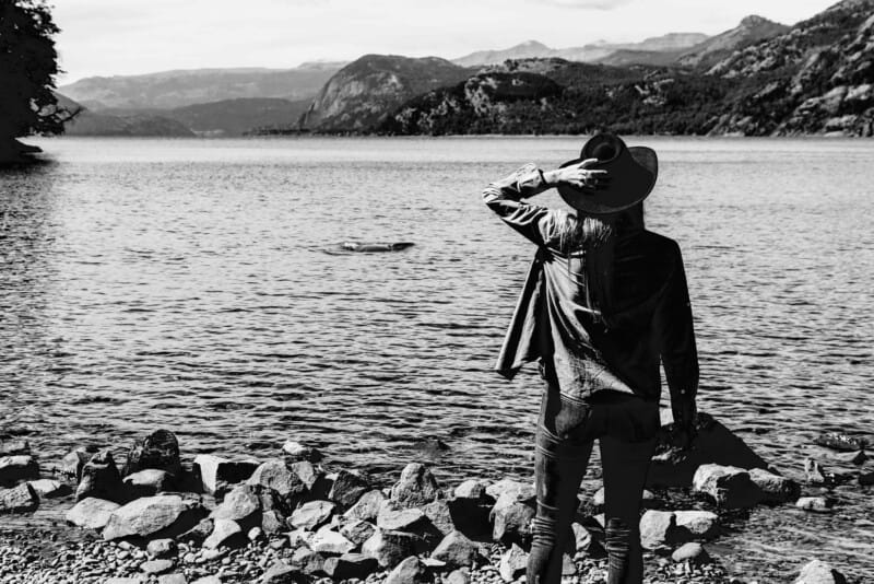 A black and white image of a woman with her back to the camera in front of a lake