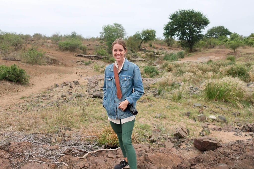 A woman in a jean jacket holds a camera and poses in Kruger National Park