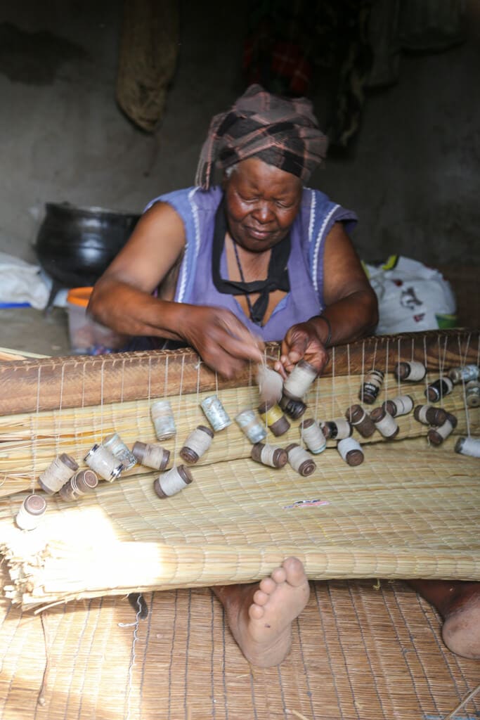 A woman weaves a grass mat in her lap