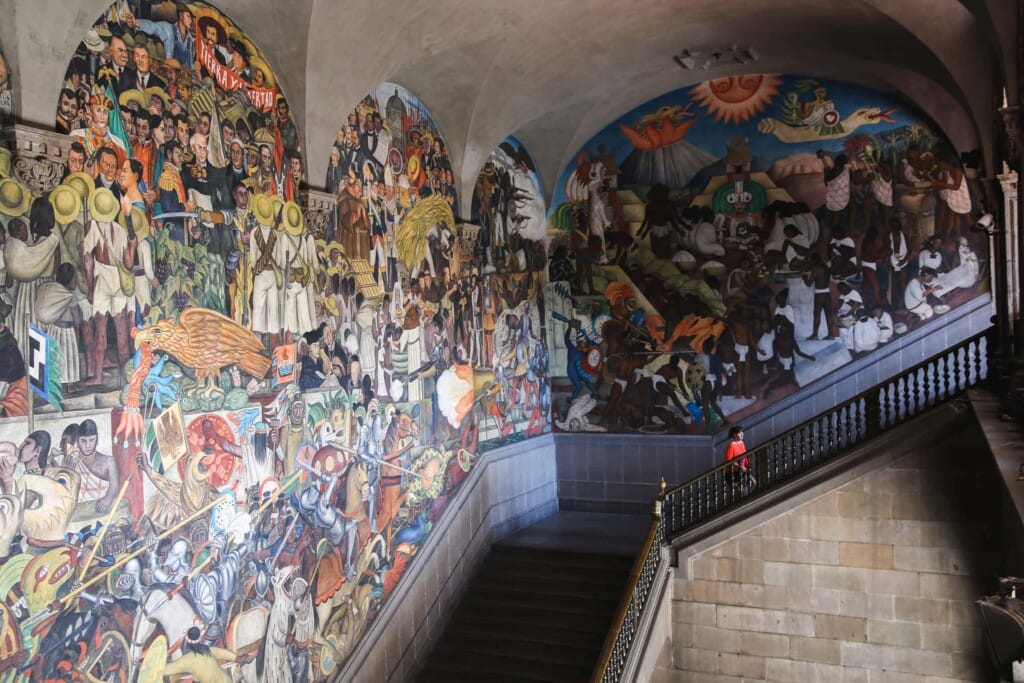 A woman in an orange shirt walks up the stairs in front of Diego Rivera murals in Mexico City