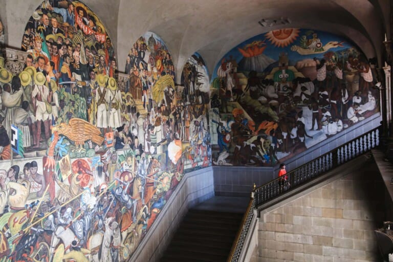 A woman in an orange shirt walks up the stairs in front of Diego Rivera murals in Mexico City