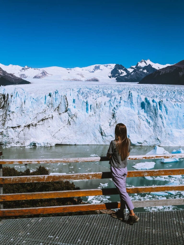 A woman stands at a handrail looking at a glacier