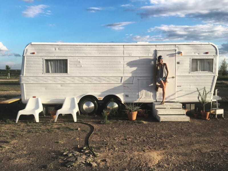 A woman stands in front of a white trailer in the desert