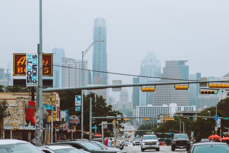 Cars drive down a busy street with the city skyline in the background