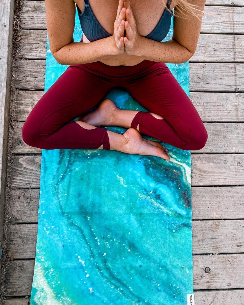 A woman sitting a top a travel yoga mat with hands in prayer at her heart
