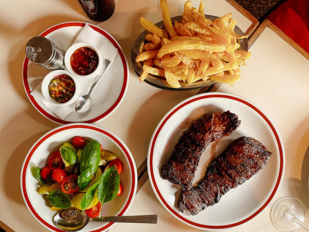 A white table with four plates, one with two steaks, one with tomatoes and basil, another with fries and a fourth with chimichurri