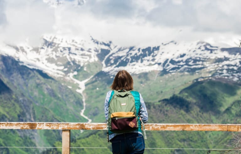 A woman in a blue shirt and jeans and a green backpack is seen from behind as she leans against a guard rail with a view of snow covered mountains in the background