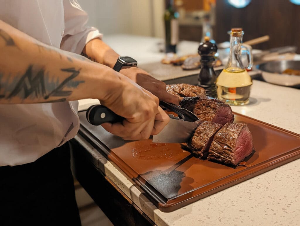 A man holds a chef's knife cutting into a tender steak on a brown cutting board