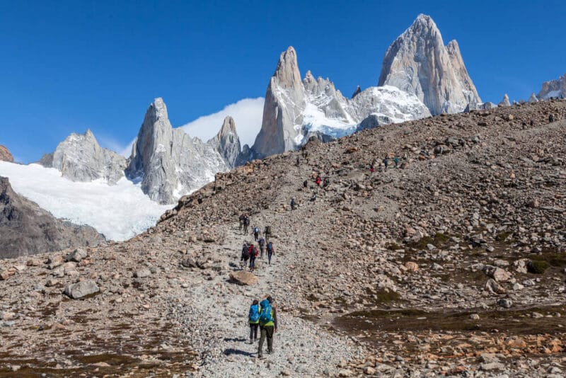 A trail of hikers are seen walking up a rocky trail with the peaks of Chalten in the background