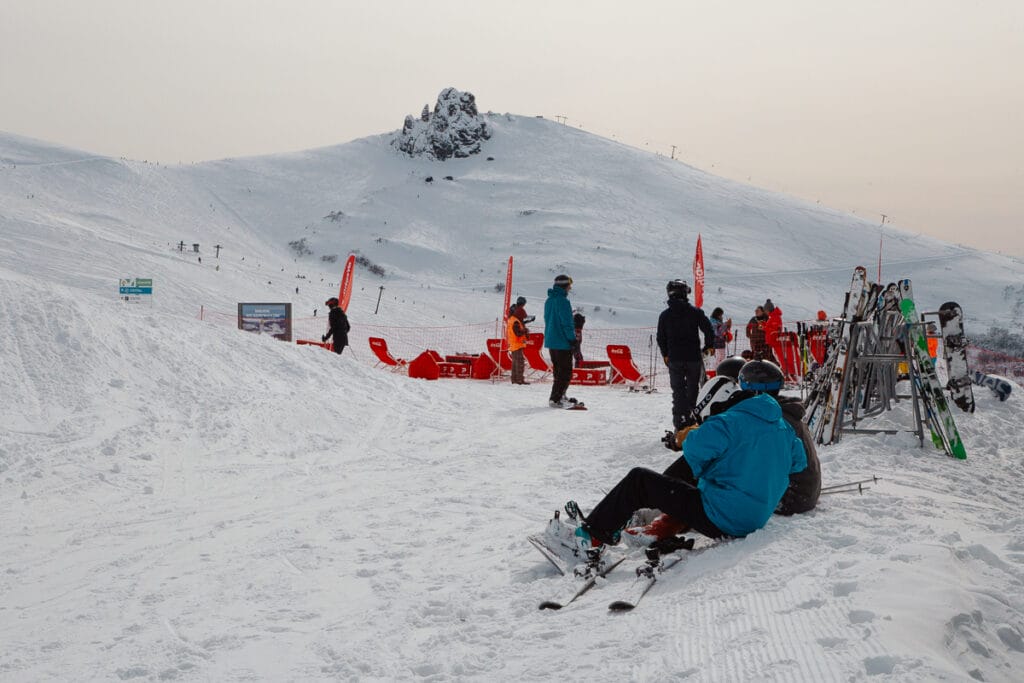 Skiers and snowboarders sit in the snow eating lunch with their skis leaning against a rack at an Argentina ski resort in Bariloche