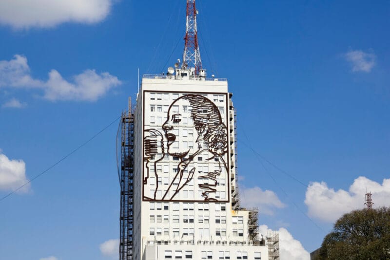 A metal portrait of Evita Peron is attached to a white office building with a blue sky in the background