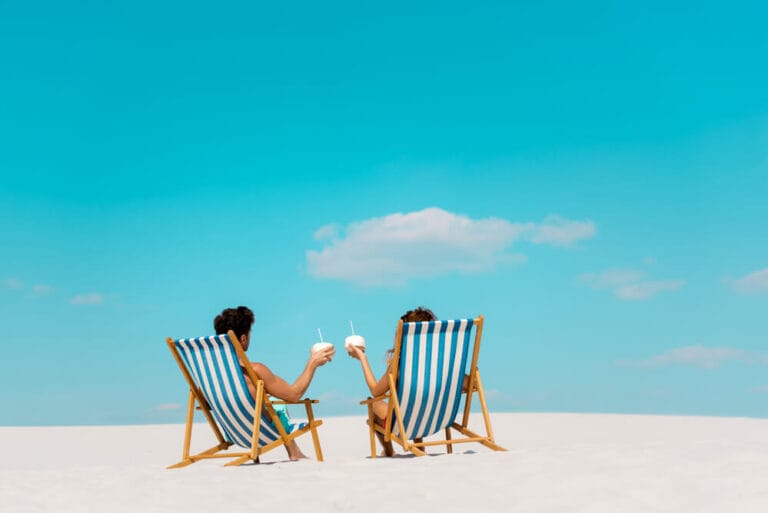 A couple sit in white and blue striped lounge chairs on a sandy beach holding white fruity cocktails in front of a bright blue sky