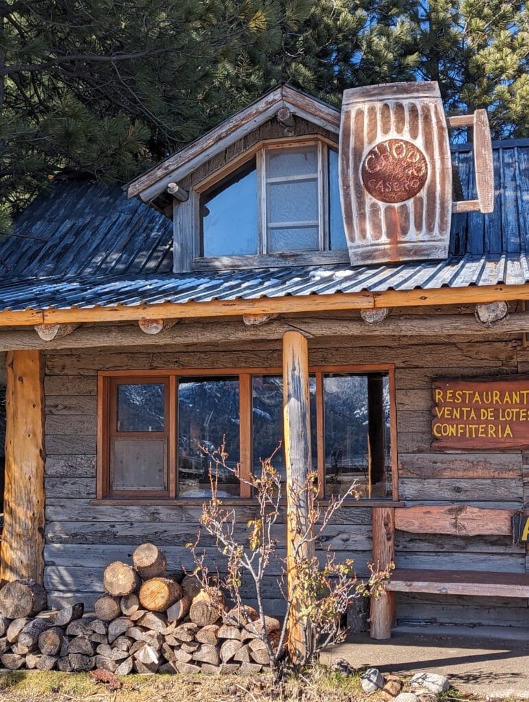 A pile of logs sits on the ground in front of the porch of a log cabin restaurant with a wooden pint of beer on the roof as the sign