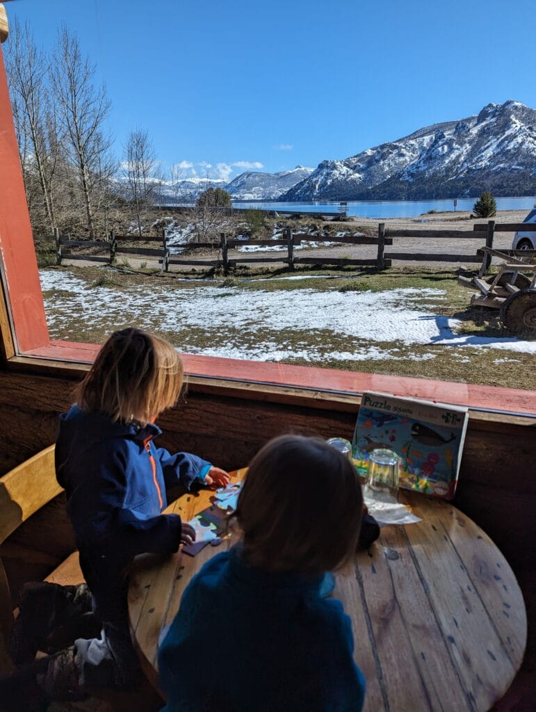 Two toddlers sit at a wooden table in front of a window with an epic view of Lago Meliquina and snow covered mountains near San Martin de los Andes