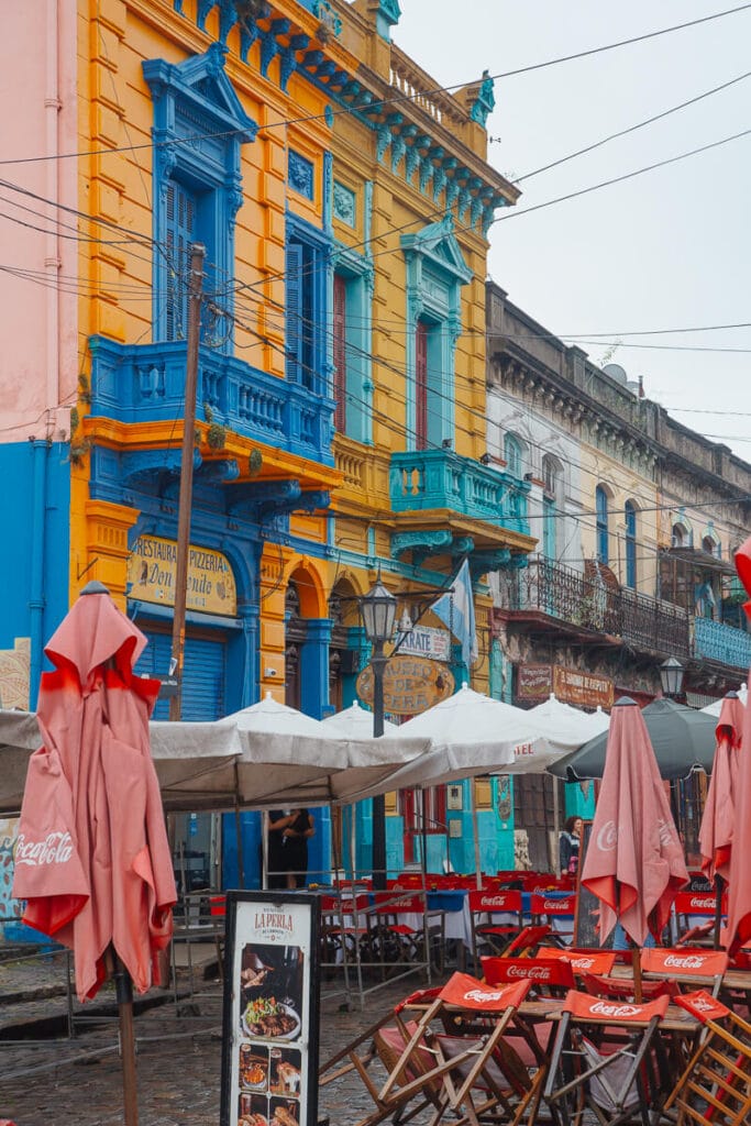 Red chairs lean against wooden tables and closed umbrellas in front of yellow and blue buildings in colorful Buenos Aires' La Boca on a rainy day