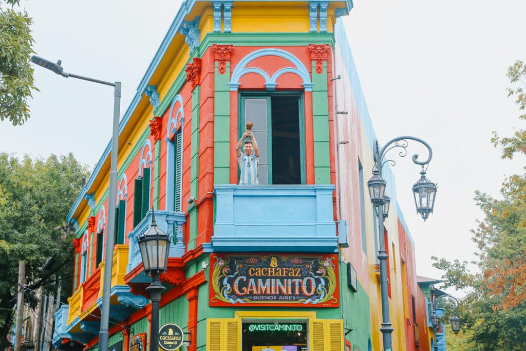 A statue of Lionel Messi holding the world cup into the air on a balcony of a rainbow colored building in the Caminito of Buenos Aires