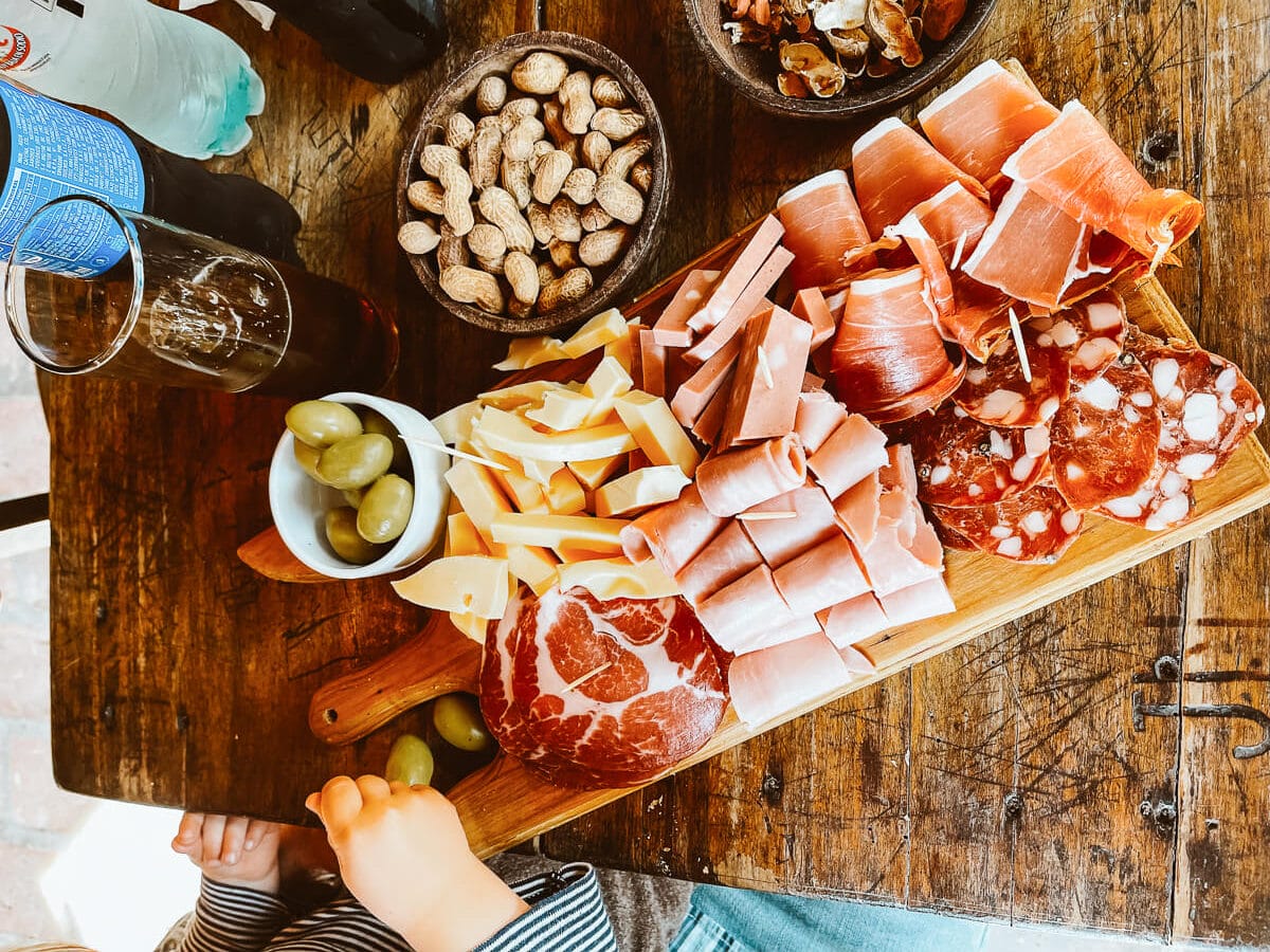 A platter of salami, hams and cheese on a wooden table with olives, peanuts, and a soda