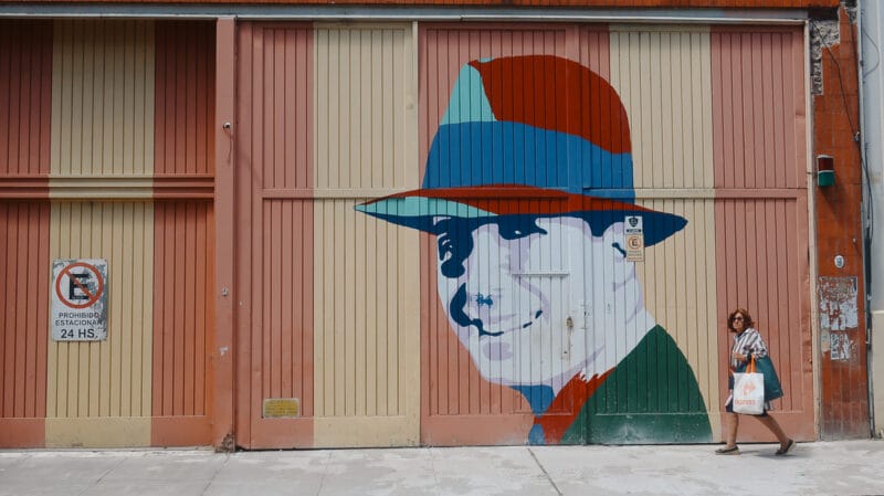 A woman walks on a sidewalk in front of a mural of Carlos Gardel