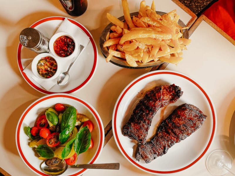 Two steaks on a red rimmed plate next to a metal dish of french fries and a fresh tomato salad at a Buenos Aires steakhouse