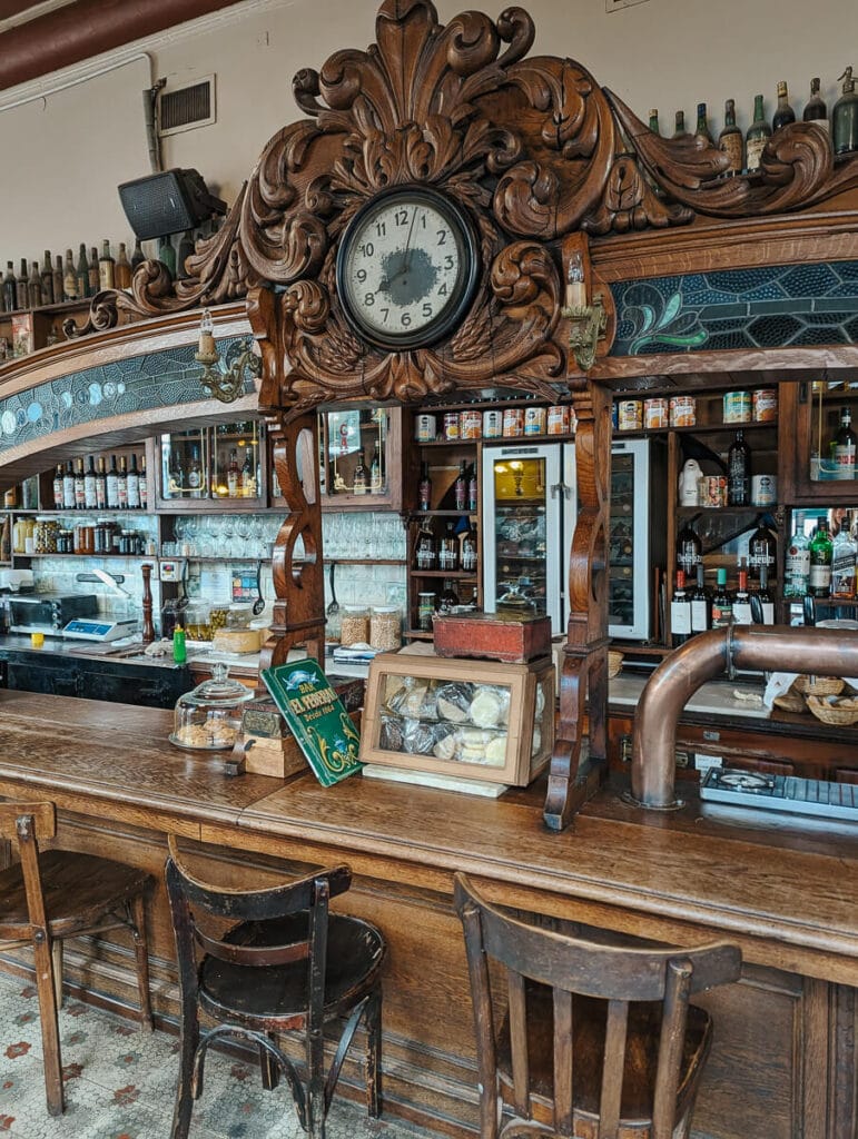 An intricately carved wooden bar with floral swirls around a clock and detailed glasswork in front of a wall filled in liquor bottles in a historic cafe in Buenos Aires
