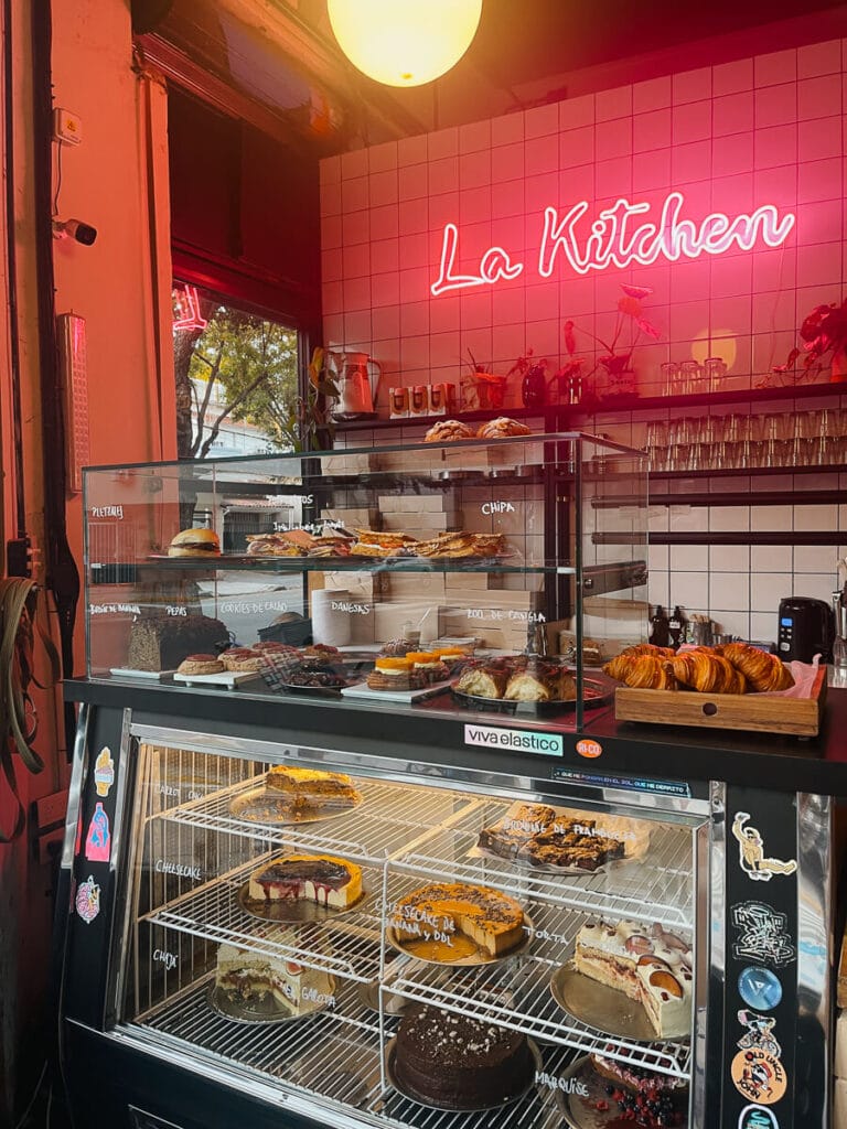 La Kitchen shines in bright pink neon lights against white tiles behind a display case full of cakes and pastries at a Buenos Aires cafe