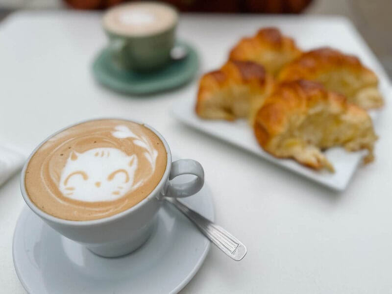 Four gooey medialunas on a white plate by two plates of coffee with latte art of a happy kitten