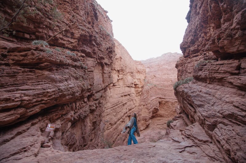 A woman in jeans and a poncho stands atop a red earth canyon with stone walls towering up on both sides of her